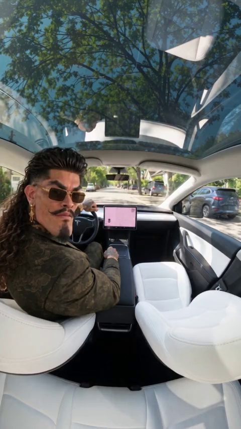Frame from the Day 1 of Owning a Roadster AI video scene: a man in sunglasses and a patterned jacket glances back from the driver seat of an electric car with white seats and a glass roof